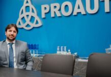 PROAUTO chega ao Dia do Corretor com modelo de parceria baseado em receita recorrente Homem de terno cinza sentado em sala de reunião diante da parede azul com o logotipo da PROAUTO